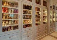 Chic walk-in closet features a wall of floor to ceiling cabinets fitted with glass front doors showcasing tilted shoe shelves lit by custom lighting.