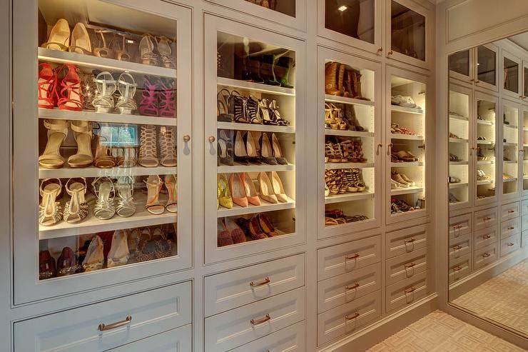 Chic walk-in closet features a wall of floor to ceiling cabinets fitted with glass front doors showcasing tilted shoe shelves lit by custom lighting.