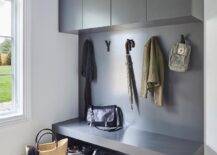 A dark gray mudroom bench with a built-in shoe shelf is fixed against a gray backsplash and under gray flat front cabinets.
