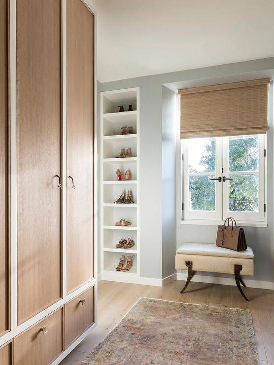 A bamboo roman shade hangs covering casement windows over a white bench placed beside inset white shoe shelves. The closet also boasts white and beige wardrobe cabinet doors with faux bois hardware.
