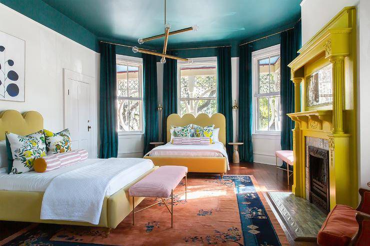 Contemporary bedroom features a yellow painted fireplace mantel with mosaic tiles, a yellow scalloped headboard with lemon print pillows and a pink stripe bolster pillows under a bay window with peacock blue velvet curtains, a matching bed under art and a peacock blue painted ceiling.