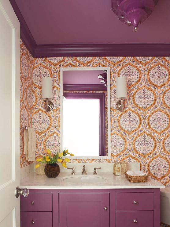 Stylish purple and orange powder room boasts walls covered in purple and orange wallpaper lined with purple crown moldings complementing a purple painted ceiling. A white vanity mirror is mounted between sconces and over a purple wooden washstand finished with a white marble countertop.
