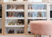 A pink velvet stool is placed in a custom closet in front of gold framed glass front shoe cabinets.