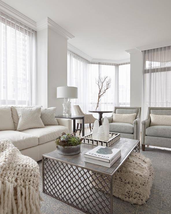 Light themed transitional living room cozies up with an ivory sofa paired with gray accent chairs around a wooden and metal coffee table. A morrocan wedding blanket ottoman under the coffee table boasts texture adding excitement to the simple yet decorated living space.