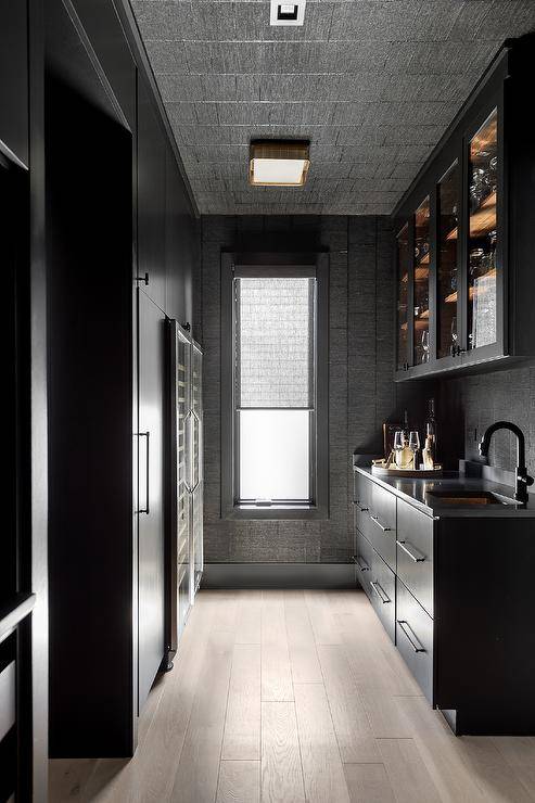 Beautiful galley style modern kitchen pantry features matte black flat front cabinets donning oil rubbed bronze pulls and a black countertop holding a sink beneath a matte black gooseneck faucet. The faucet is fixed beneath glass front cabinets mounted to black textured wall tiles under a black textured tiled ceiling.