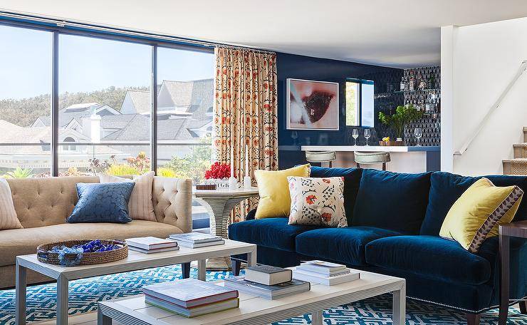 A jewel blue velvet sofa is complemented with bright yellow pillows and sits on a white and blue geometric rug facings side-by-side coffee tables.