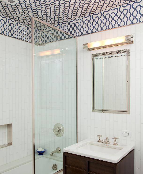 Under a white and blue wallpapered ceiling, an inset framed medicine cabinet is fixed to white vertical wall tiles over a brown wooden washstand finished with a vintage cross handle faucet mounted to a marble countertop. A drop in bathtub with a glass partition is located beside the washstand and completed with white vertical surround tiles.
