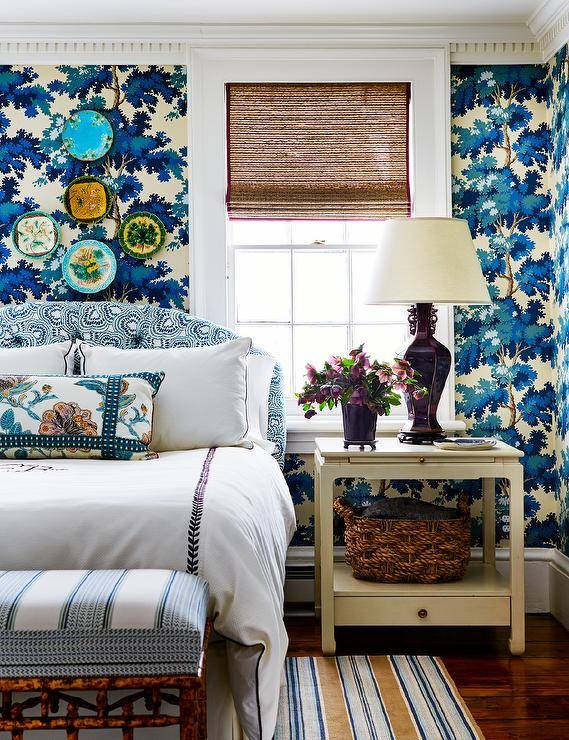 Bedroom features a white and blue headbaord on cream and blue Raphael Sandberg wallpaper, a cream chinoiserie nightstand lit by a burgundy lamp and a blue and beige striped rug.