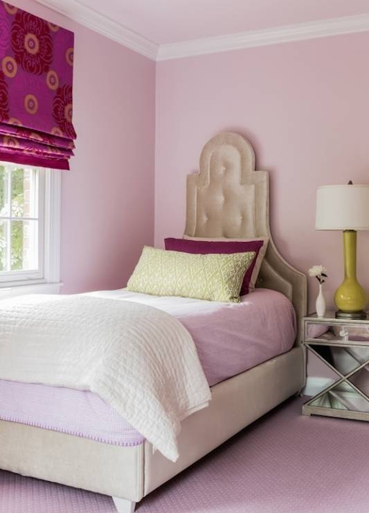 Chic girl's room with pink ceiling over pink walls framing an arched beige velvet tufted headboard on bed layered with pink and ivory bedding topped with a burgundy sham and green diamond print lumbar pillow. An x-base mirrored nightstand stands beside the bed, topped with a chartreuse table lamp across from the bedroom window which is dressed in a fuchsia and burgundy floral print shade.