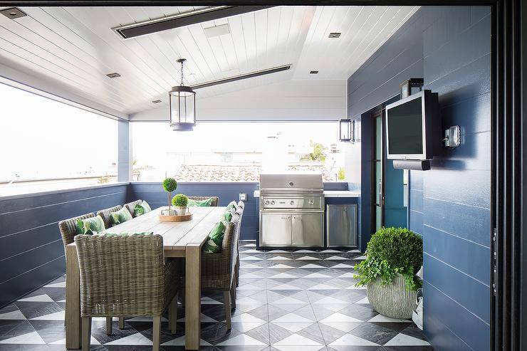 An oil rubbed bronze and glass lantern is hung in a covered patio from a white shiplap ceiling over a teak slatted dining table placed on black and white triangle pattern floor tiles and surrounded by wicker dining chairs. A television is mounted to a glossy dark blue shiplap wall, while the patio is equipped with a stainless steel barbecue.