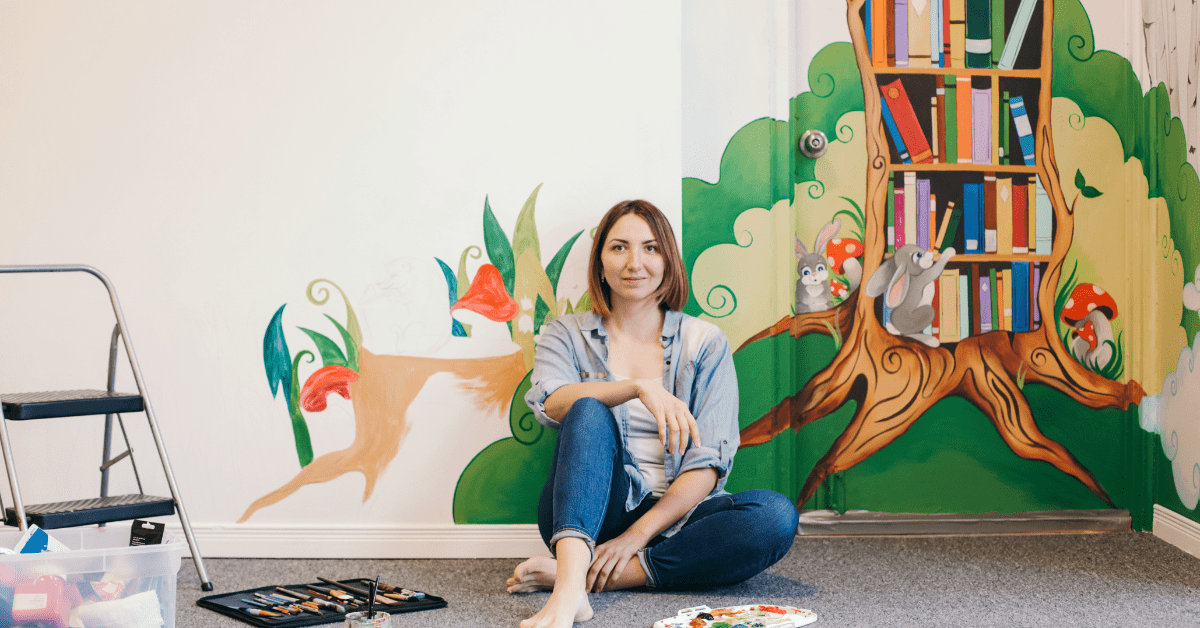 Woman with paint in front flanked by a wall of kids room art that she painted.