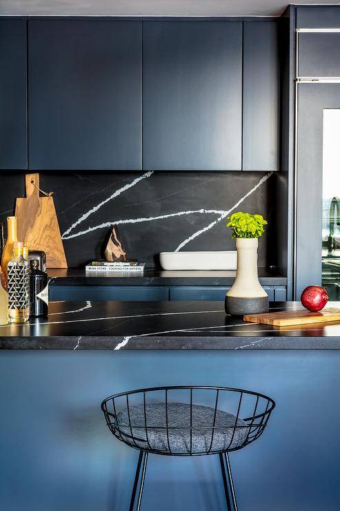 Blue and black chic contemporary kitchen features low back black metal counters stools placed at a blue island topped with a honed black marble countertop. Behind the island, blue flat front cabinets with a honed black marble countertop are mounted against a honed black marble backsplash beneath blue flat front upper cabinets.