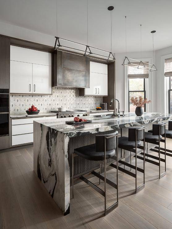 Kitchen features a black and white waterfall edge island with metal trim and black and gold stools lit by dual linear island lights, a custom range hood with hidden TV over white gray black mosaic backsplash tiles and white and gray cabinets.