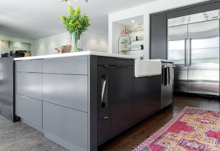 A pink runner sits on an stained oak wood floor at a charcoal gray kitchen island boasting modern flat front cabinets, a stainless steel dishwasher and a farmhouse sink with a satin nickel gooseneck faucet mounted to a white quartz countertop. To the side of the island, a Viking refrigerator is inset between charcoal gray flat front cabinets.
