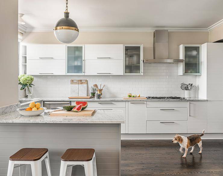 Kitchen features white metal and wood Tolix stools at a granite top peninsual lit by a Hicks pendant and white cabinets on white subway tiles.