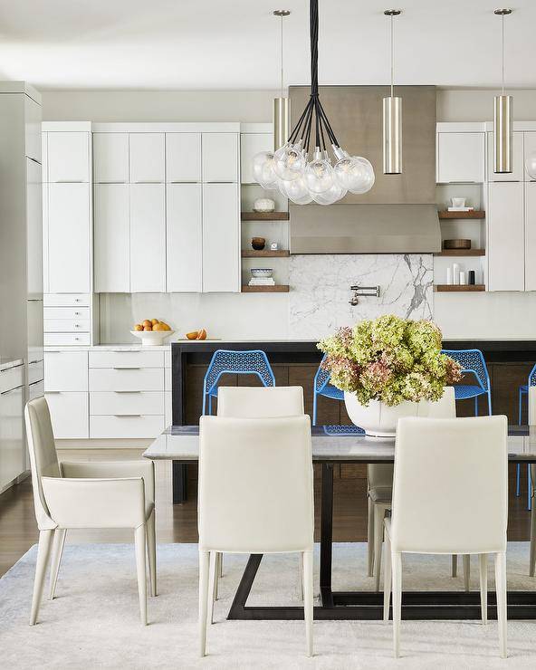 Modern kitchen design features off white leather chairs at a gray marble and black metal dining table lit by a modern light bulbs chandelier and white cabinets with nickel pulls.
