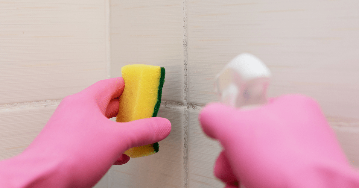 Closeup of someone's hands in pink gloves cleaning tile grout with sponge and spray bottle.