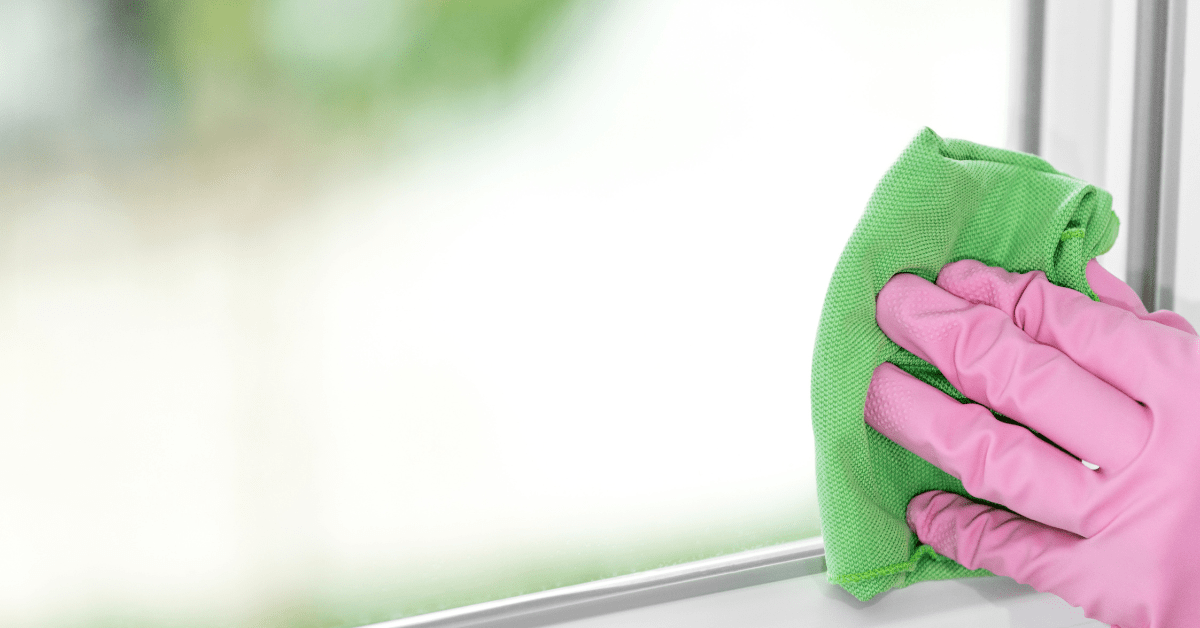 Closeup of person's hand wearing pink gloves using a green microfiber to clean window.