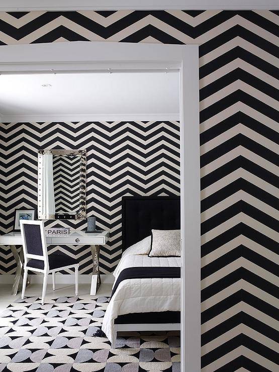 Contemporary black and white bedroom features black and white chevron wallpaper, a black and white French bed, a chrome x base desk as nightstand under a studded mirror with a black and white French chair and a black, white and gray rug.