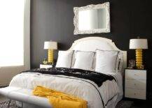 Black and white bedroom with yellow accents features a black accent wall holding a white baroque mirror over a white French headboard. The bed is dressed in white and black bedding complemented with black and white Greek key pillows, and it sits on a black animal print rug behind a white leather bench. The bed is positioned between black and white nightstands complemented with yellow lamps.
