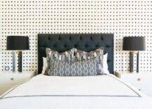 Black and white wallpaper complements a black tufted headboard accenting a bed dressed in black border hotel bedding topped with black and gray pillows. The bed is flanked by white lacquer nightstands illuminated by black lamps.