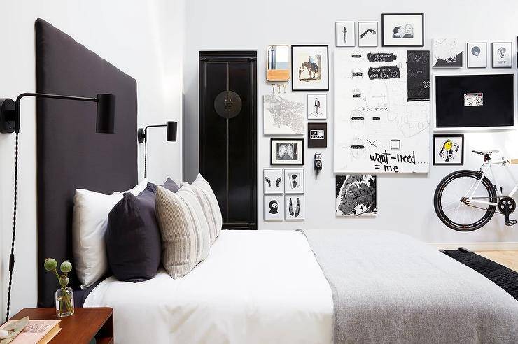 Chic black and white contemporary bedroom boasts a tall black headboard lit by black plug in sconces and accenting a bed dressed in white and gray bedding topped with gray and black pillows. A tall black armoire is located beside a gorgeous black and white gallery art wall.