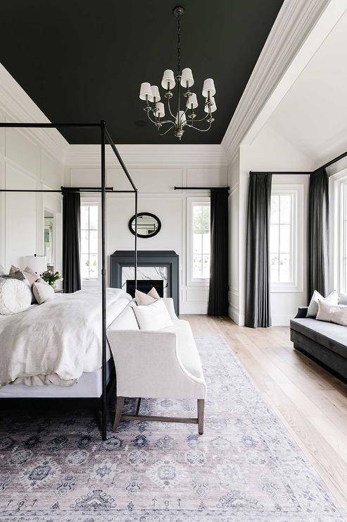 Black and white bedroom features a white settee at the foot of a black canopy bed with white headboard accented with pink and gray velvet pillows, a black ceiling, white wall trim, black curtains and a vintage rug.