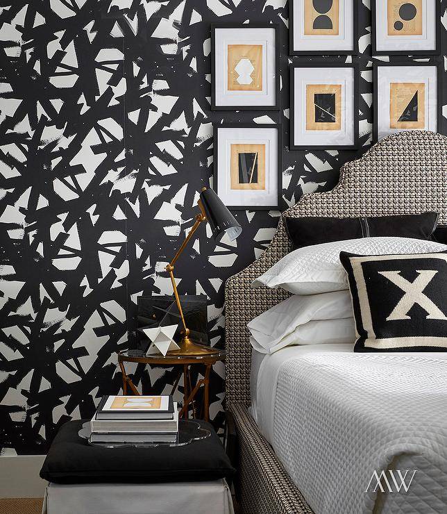 Tan and black in abstract art in black frames hangs from a wall covered in black and white wallpaper over a beige and black houndstooth headboard. The bed is topped with white and black pillows placed on white waffle weave bedding. A black and gold task lamp sits on a gold bedside table beside the bed.