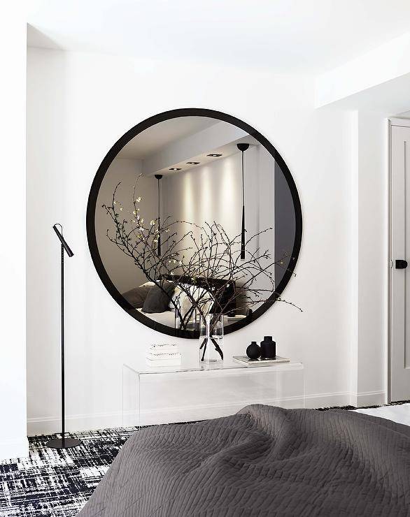 An extra large round black mirror hangs over a lucite bedroom bench placed on a black and white rug lit by a black floor lamp.