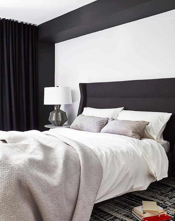 A gray lamp lights a round marble top bedside table placed on a black and white rug beside a black wingback headboard accented with a gray chevron quilt and gray pillows. The bed is placed against in a black and white alcove, while a window is covered in black curtains.