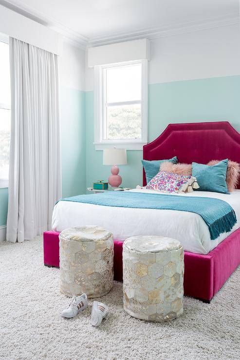 Blue velvet pillows decorate a fuchsia velvet tufted bed in a girls bedroom designed with pink double gourd lamps on nightstands. An aqua blue wall contrast the bold fuchsia headboard with the perfect balance while gray and gold hexagon stools accent the foot of the bed.