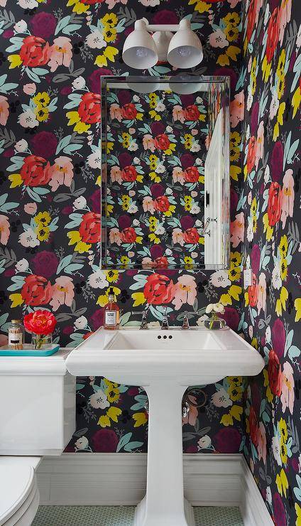 Covered in bold floral wallpaper, this welcoming contemporary powder room features a white pedestal sink fitted with a nickel faucet and fixed to green penny tiles beneath polished nickel mirror lit by a white two light sconce. An aqua tray sits atop a the white porcelain toilet located beside the sink vanity.