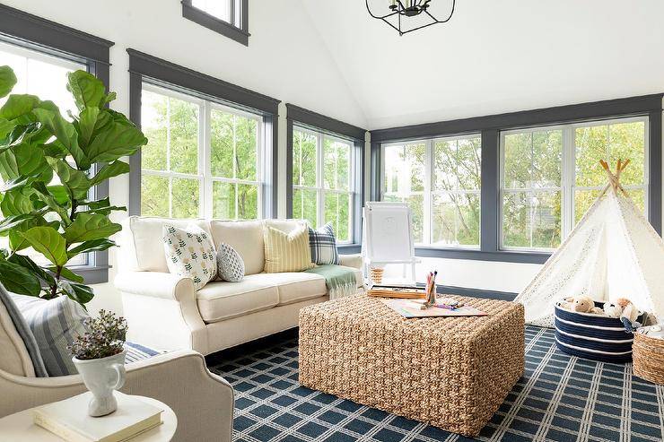 Rectangular woven coffee table in a sunroom designed with a white roll arm sofa, a blue check rug, and a fiddle leaf fig plant. Blue window moldings accent the windows and bring a contrast to white walls and natural woven accents.