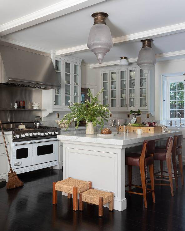 A stainless steel range hood is fixed to a stainless steel cooktop over a stainless steel cooktop rack mounted between glass front cabinets and above a white Viking stove placed on an espresso wood floor.
