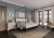 Bedroom features a black canopy bed with gold lumbar pillow flanked by light gray French nightstands lit by brass sconces, a brown rug atop parquet wood floors and blue wall panels with cream curtains.