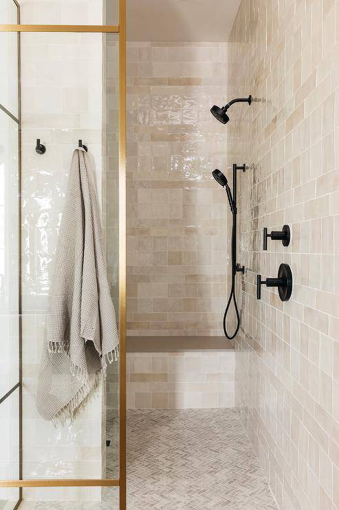 Shower features a matte black shower kit on beige glazed grid tiles with built in shower bench, gray marble mini herringbone shower floor tiles and a glass and brass shower partition.