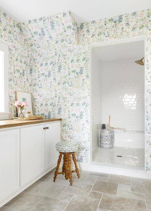 Clad in floral wallpaper, this welcoming bathroom features a doorless walk-in shower covered in white subway surround tiles and fitted with a subway tiled bench fixed behind a white and blue pierced drum stool. A wooden stool sits outside of the shower and in front of white cabinets topped with a brown wood countertop mounted beneath a window.
