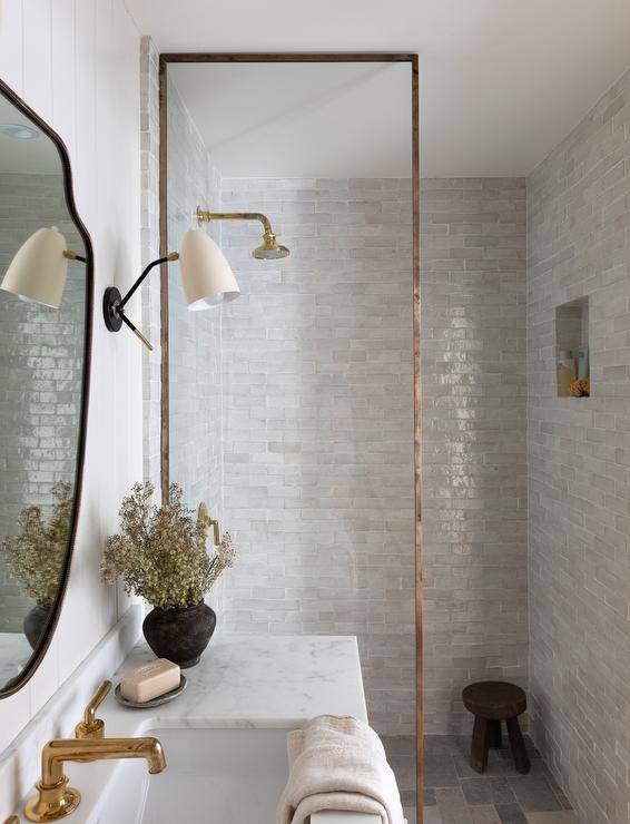 A glass partition with brass trim accents a shower clad in gray glazed surround tiles framing a niche located over a black stool. The shower is finished with a brass shower kit.