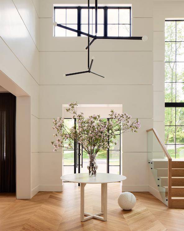 Foyer features a round accent table decorated with a clear vase of flowers and wood chevron floors lit by a modern black and white chandelier.