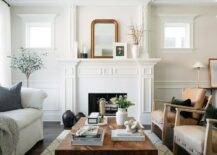 Welcoming living room boasts a white traditional fireplace styled with a gold beaded arch mirror. A burl wood coffee table sits between light brown leather chairs and a natural linen sofa.