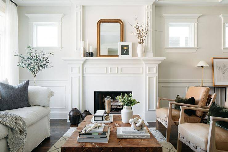Welcoming living room boasts a white traditional fireplace styled with a gold beaded arch mirror. A burl wood coffee table sits between light brown leather chairs and a natural linen sofa.