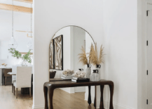 A stunning carved wood console table sits in a foyer in front of a gold arched mirror and on a wood floor.