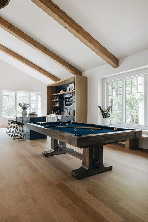 Open plan living space features a brown and blue pool table placed on a wood floor beneath a vaulted ceiling finished with wood beams.