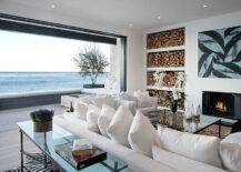 Stunning beach house with pocket patio doors opening onto incredible deck with wraparound bench and frameless glass railing providing unobstructed views of the ocean. The living area features a large abstract mounted above a minimalist style fireplace with stacked firewood alcoves to the left. A pair of white slipcovered sofas face the fireplace with an iron and glass coffee table in front and an iron and glass sofa table behind atop faux zebra hide rugs layered over hardwood floors.