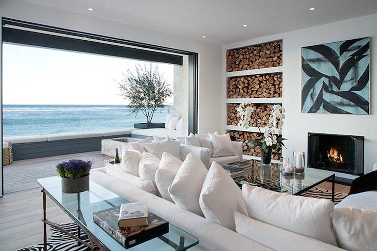 Stunning beach house with pocket patio doors opening onto incredible deck with wraparound bench and frameless glass railing providing unobstructed views of the ocean. The living area features a large abstract mounted above a minimalist style fireplace with stacked firewood alcoves to the left. A pair of white slipcovered sofas face the fireplace with an iron and glass coffee table in front and an iron and glass sofa table behind atop faux zebra hide rugs layered over hardwood floors.