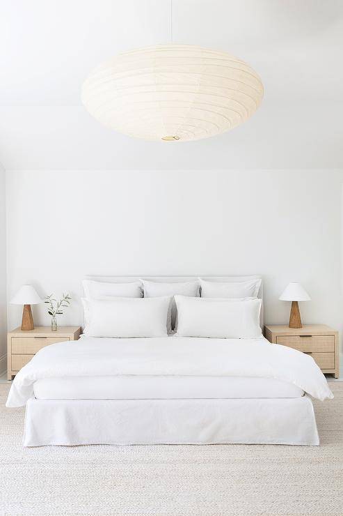 Minimalist bedroom features a white slipcovered bed accented with a white duvet and stacked white pillows and flanked by tan nightstands topped with wooden lamps. The room is illuminated by a paper chandelier.