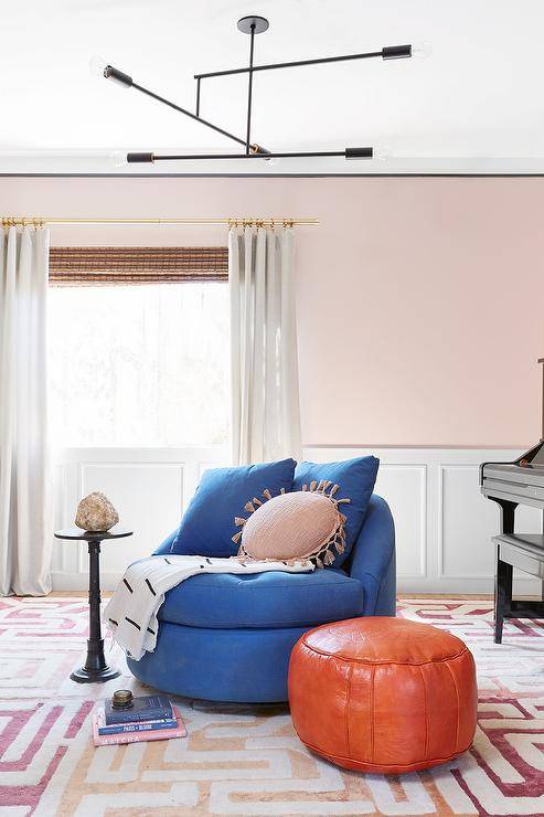Elegant contemporary playroom is accented with blush pink walls lined with wainscoting and complementing white curtains hung from a brass rod in front of a bamboo roman shade. A piano sits in a corner behind a round blue lounge chair placed on a colorful geometric rug and matched with an orange leather Moroccan pouf lit by a minimalist contemporary chandelier.