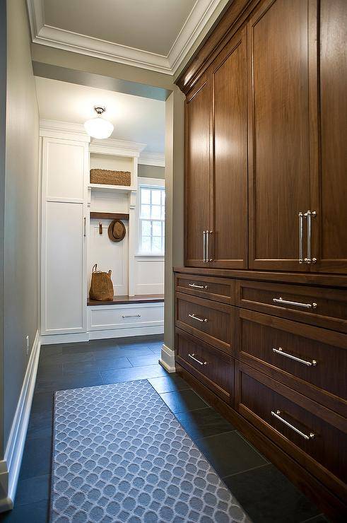 Tall floor to ceiling, brown stained mudroom cabinets, make the most of this hall space allowing storage to be concealed and a built in to showcase. A gray geometric runner on slate floor tiles, lead into a wooden mudroom built-in bench in white. A schoolhouse pendant light distinguishes two different spaces and decor schemes while sharing the same floor base.