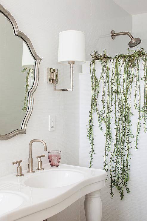 A silver leaf mirror flanked by sconces is mounted over a French pedestal dual sink vanity finished with satin nickel faucets. Plants sit atop a shower partition fixed beside the vanity.