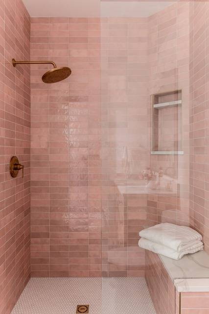 Shower features pink horizontal stacked tiles with an antique brass shower kit and niche, a built in marble slab shower bench and white hexagon shower floor tiles.